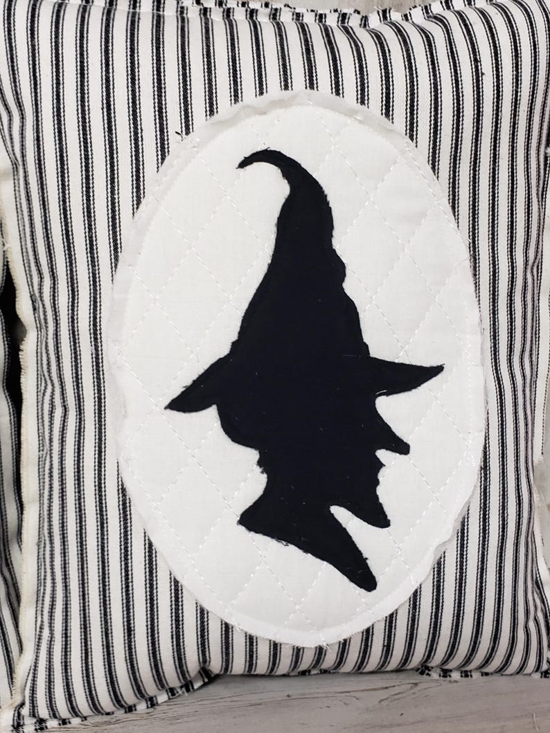 May include: Black and white striped pillow with a white oval patch featuring a black silhouette of a witch wearing a pointed hat.