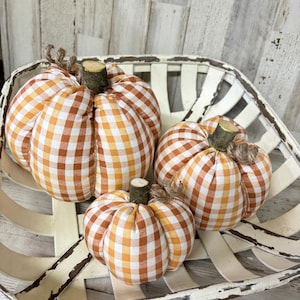 May include: Three fabric pumpkins in a white basket. The pumpkins are made of orange and white checked fabric, with wooden stems and twine accents. The basket has a distressed, rustic appearance.