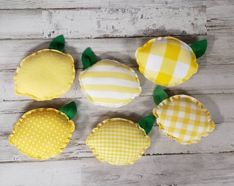 May include: Six yellow lemon-shaped pillows with green stems. The pillows are made of fabric with different patterns, including polka dots, stripes, and gingham.