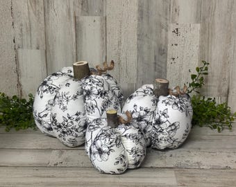 Könnte beinhalten: Vier Stoffkürbisse in verschiedenen Größen, verziert mit einem schwarzen Blumenmuster auf weißem Grund. Jeder Kürbis hat einen Holzstiel und Bindfaden-Akzente, ideal für die Herbstdekoration. Die Kürbisse sind auf einer Holzoberfläche angeordnet.