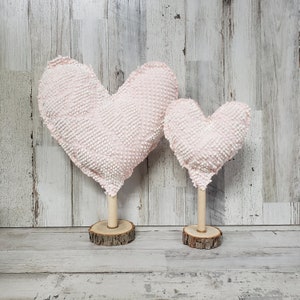 May include: Two pink heart-shaped pillows with white polka dots, standing on wooden bases. The larger heart is in the background and the smaller heart is in the foreground.