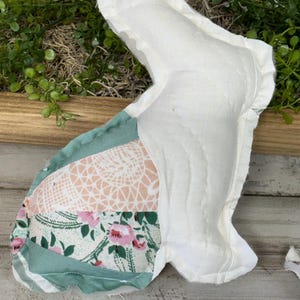 May include: A handmade, stuffed bunny-shaped decoration. The bunny features a white body and patchwork ears with floral and lace patterns in pink, green, and white. The bunny is placed on a wooden surface.