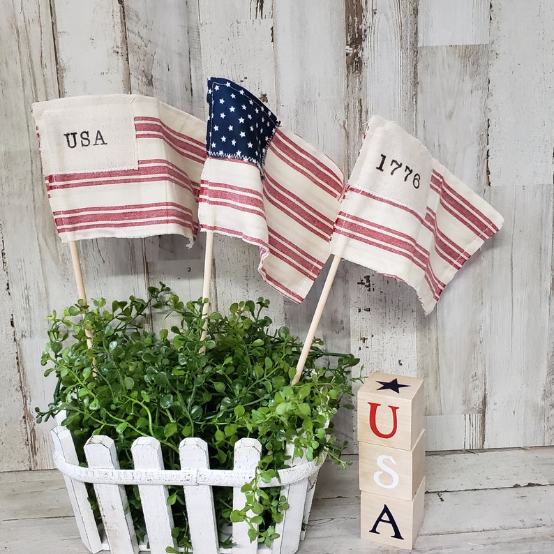May include: Three miniature American flags with red and white stripes and blue stars, displayed in a white picket fence planter with green foliage. The flags are labeled "USA", "1776", and a star.