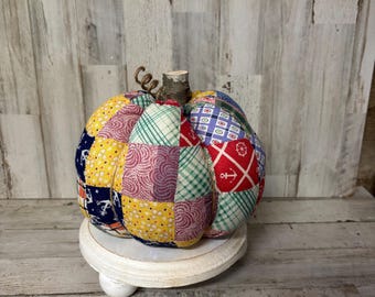 Può includere: Una zucca in tessuto fatta a mano, con un design patchwork nei toni del giallo, blu navy, rosso e verde. Ha un gambo di legno e un viticcio. La zucca è esposta su un piccolo piedistallo rotondo bianco, ideale per l'arredamento autunnale.