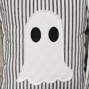 May include: A white ghost pillow with black eyes on a black and white striped fabric background.