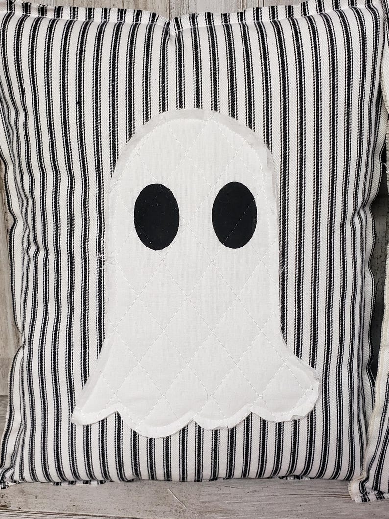 May include: A white ghost pillow with black eyes on a black and white striped fabric background.
