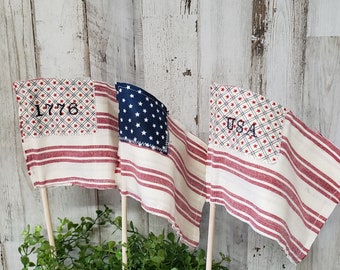 May include: Three small American flag bunting banners with red and white stripes and a blue star field. The banners are made of fabric and have the text "1776", "USA", and a blue star field on them. The banners are hanging from wooden sticks.
