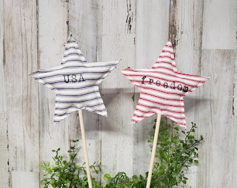 May include: Two fabric stars on sticks, one red and white with "freedom" written on it, and one blue and white with "USA" written on it. The stars are sitting in a pot of green plants.