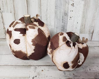 May include: Two brown and white patterned fabric pumpkins with twig stems. The pumpkins are sitting on a whitewashed wood surface.