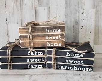 Op de afbeelding: Drie sets gestapelde houten blokken met zwarte en bruine verf. De blokken zijn vastgebonden met touw en hebben witte letters. De bovenste set luidt "home sweet home", de middelste set luidt "farm sweet farm" en de onderste set luidt "home sweet farmhouse".