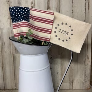 May include: A white metal pitcher with a black rim and handle holds a small American flag and a cream-colored fabric with the text "1776" and stars. The flag has a blue field with white stars and red and white stripes.