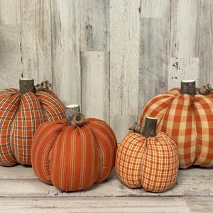 May include: Four fabric pumpkins in varying sizes and patterns, including orange plaid, striped, and gingham designs. Each pumpkin features a textured wooden stem at the top, creating a collection of soft, stuffed decorative gourds.