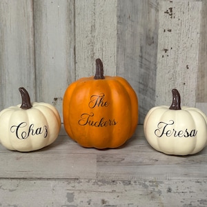 May include: Three decorative pumpkins with brown stems. The center pumpkin is orange and has "The Tuckers" written on it. The pumpkins on either side are white and have the names "Chaz" and "Teresa" written on them.
