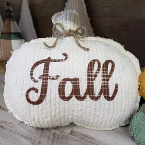 May include: A white, plush pumpkin decoration with the word "Fall" in brown plaid fabric. The pumpkin has a stem made of the same material and is tied with twine. Other decorative items are visible in the background.