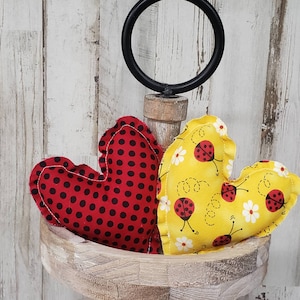 May include: Two fabric hearts with a ladybug print. One heart is red with black polka dots and the other is yellow with white flowers and black ladybugs. The hearts are sitting on a wooden tiered stand.
