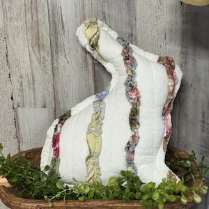 Peut inclure: Un coussin en forme de lapin blanc et matelassé, avec des accents verticaux en tissu floral. Le lapin est dans un bol en bois avec de la verdure, créant un thème de Pâques ou de printemps rustique. Le design présente un mélange de couleurs pastel.