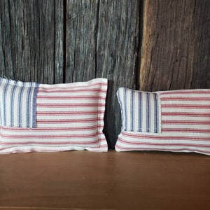 May include: Two small decorative pillows in the shape of the American flag. The pillows are made of a white fabric with red and blue stripes. The pillows are sitting on a wooden surface.