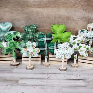 May include: A collection of nine green shamrock-shaped decorations with various patterns, including stripes, polka dots, and shamrock designs. Each shamrock is attached to a wooden stick and placed on a wooden base.