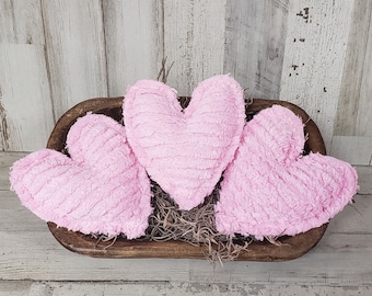 May include: Three pink, heart-shaped decorative pillows with a textured surface, arranged in a wooden bowl filled with dried moss. The bowl has a dark brown finish, and the background is a distressed white wood.