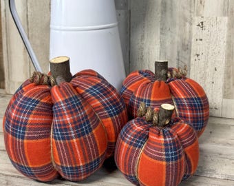 May include: Three fabric pumpkins featuring an orange, navy blue, and white plaid pattern. Each pumpkin has a natural wooden twig stem and is wrapped with thin jute twine. The pumpkins are arranged in a group with a white metal pitcher behind them.