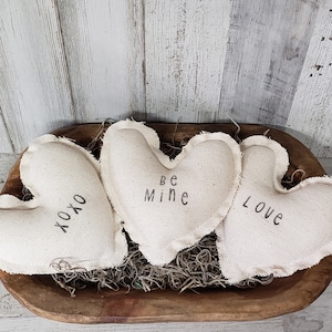 以下が含まれることがあります： 「XOXO」、「Be Mine」、「Love」と印刷された白い布製のハート型のクッションが3つ。クッションは、茶色のわらで満たされた木製のボウルに入っています。