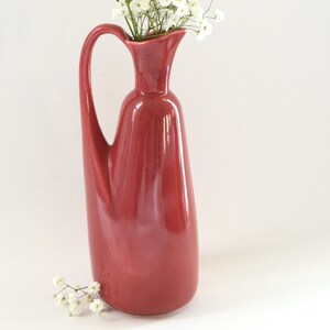 May include: A tall, red ceramic vase with a curved handle, filled with a bouquet of small white flowers. The vase has a slender, elegant shape and sits on a white surface. A few loose flowers are scattered near the base.