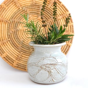 May include: A white ceramic vase with a raised floral design, filled with green leafy plants, sits in front of a round woven wicker wall decor.