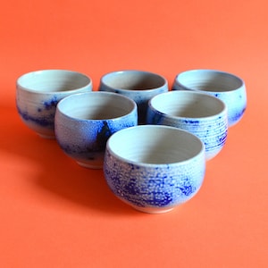 Puede incluir: Seis cuencos pequeños de cerámica con un esmalte blanco y motas azules. Los cuencos están dispuestos en círculo sobre un fondo naranja brillante.