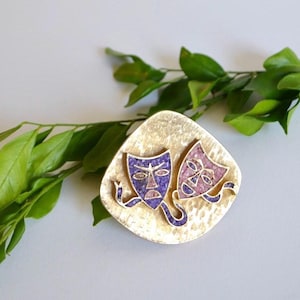 May include: A gold-toned brooch with two colorful masks. The masks are made of a textured material and are painted in shades of purple and pink. The masks are attached to a gold-toned base with a hammered texture.