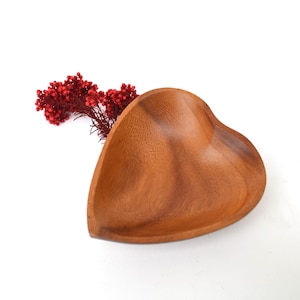 Puede incluir: Un cuenco de madera en forma de corazón con un acabado natural de vetas de madera. El cuenco es de color marrón cálido y tiene una superficie lisa. Una ramita de bayas rojas se coloca junto al cuenco.