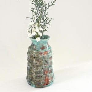 May include: A handcrafted ceramic vase with a textured, zig-zag pattern in shades of teal, brown, and copper. The vase holds a sprig of greenery and small white flowers, creating a natural, decorative display.