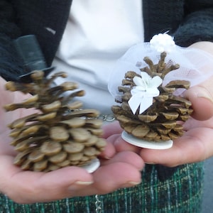 May include: Two pinecone wedding cake toppers. One pinecone has a miniature black top hat, the other has a white veil and bow. Both are on small white bases and connected by a silver chain.