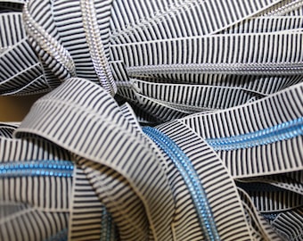 May include: Close-up of black and white striped ribbon with silver and blue zippers. The ribbon is folded and twisted, creating a textured and abstract pattern.