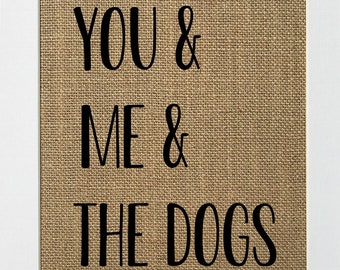 May include: A burlap sign with the text "YOU & ME & THE DOGS" printed in black.