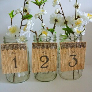 May include: Three clear glass jars with burlap table number cards. The jars are filled with white flowers and have a brown lace trim around the top. The table numbers are 1, 2, and 3.