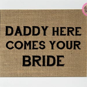 Puede incluir: Un letrero de arpillera con el texto "Daddy Here Comes Your Bride" en letras negras.