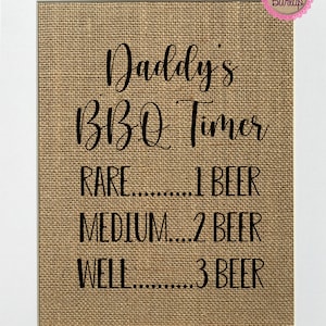 May include: A brown burlap sign with black text that reads "Daddy's BBQ Timer" and lists the doneness of meat with a corresponding number of beers. The sign is framed in white.