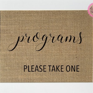 Puede incluir: Un letrero de arpillera con el texto "programs" en letra cursiva y el texto "PLEASE TAKE ONE" en mayúsculas.