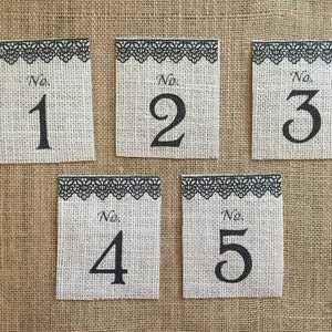 Może przedstawiać: Pięć jutowych kartek z numerami stołów z czarną koronkową lamówką i czarnymi cyframi. Numery to 1, 2, 3, 4 i 5.