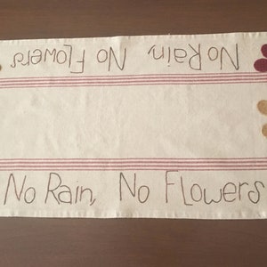 May include: A white table runner with red stripes and the text "No Rain, No Flowers" embroidered in black. The runner is decorated with four felt flowers in red, yellow, and plaid.