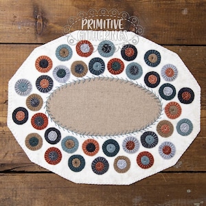 May include: An off-white, octagonal placemat with a central oval of tan fabric. The placemat is decorated with numerous small, colorful, circular appliqués in shades of orange, blue, green, and gray. The text "PRIMITIVE GATHERINGS" is arched across the top.