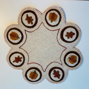 May include: A cream-colored felt rug with a scalloped edge and a brown stitched design. The rug has eight circles with brown felt pumpkins and leaves.