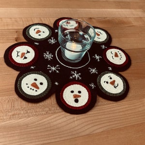 May include: A black felt snowman-themed table runner with a clear glass candle holder in the center. The runner has seven embroidered snowman faces with red noses and black eyes. The runner is surrounded by snowflakes.