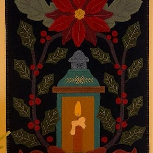 May include: A felt Christmas wall hanging with a black background. It features a red poinsettia, green holly leaves with red berries, and a teal lantern with a lit candle. A red bow is at the bottom.