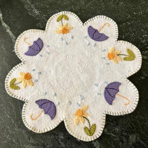Könnte beinhalten: Ein weißer Filzkreis mit einem gewellten Rand, bestickt mit violetten Regenschirmen, gelben Narzissen und grünen Blättern. Das Design erinnert an einen Frühlingsregen.