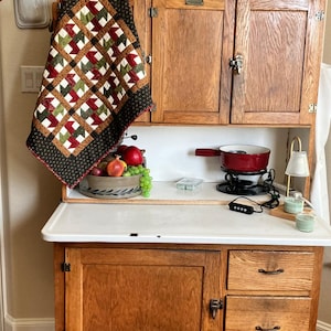 May include: A vintage wooden kitchen cabinet with a white countertop. A small, colorful quilt hangs on the side. A bowl of fruit and a red fondue pot sit on the counter. Decorative items include a ceramic hen and a vase of flowers.