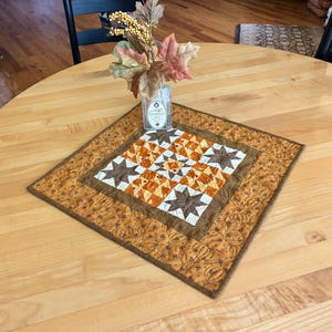 May include: A square quilted table topper featuring a central star pattern in shades of brown, orange, and cream. The border is a solid mustard fabric with a quilted pumpkin motif. A clear glass vase with artificial autumn leaves sits on top.