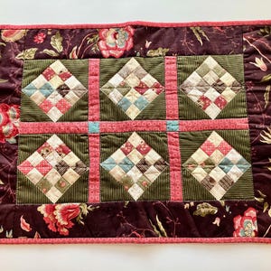 May include: A quilted placemat with a floral border and a central patchwork design. The border is a deep burgundy with floral patterns, and the center features squares of various colors, including pink, green, and cream.