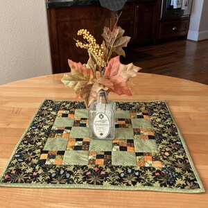 May include: A square, quilted table topper with a patchwork design in shades of green, orange, and black, with a floral border. A clear glass bottle with a label and fall foliage are on top of the topper.