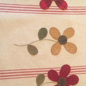 May include: A beige fabric kitchen towel with red and brown striped borders. Three appliqued flowers are stitched on the towel. The flowers are red, brown, and tan. The flowers have green leaves.